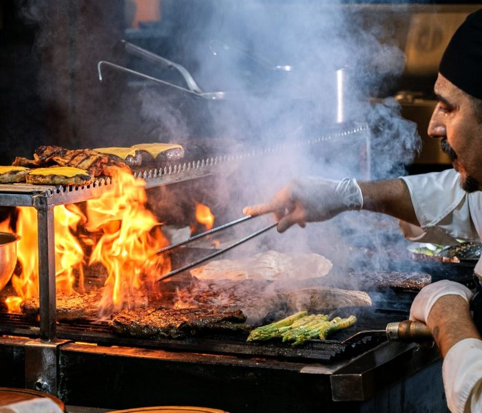 pexels-photo-36543118-36543118 A chef skillfully grills vegetables and meats over an open flame in a bustling restaurant kitchen.